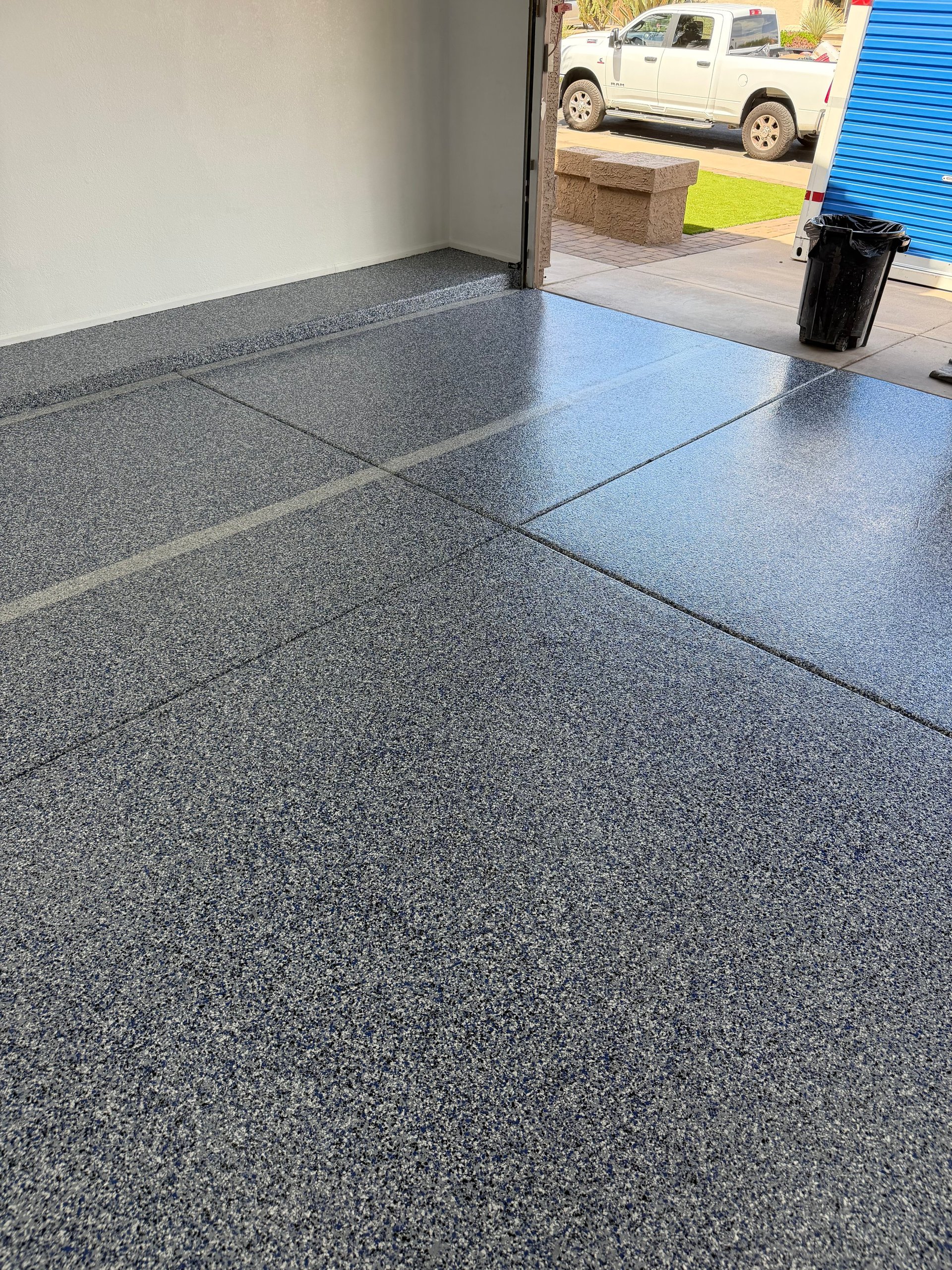 Empty garage interior with gray epoxy floor, white walls, open bay door showing parked truck and storage units outside