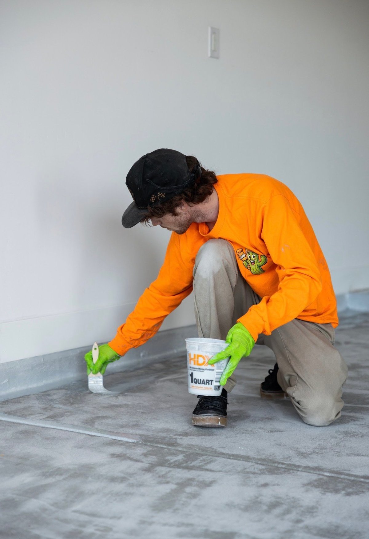 Worker in orange jacket and black cap kneeling on floor applying white paint or coating with brush and container