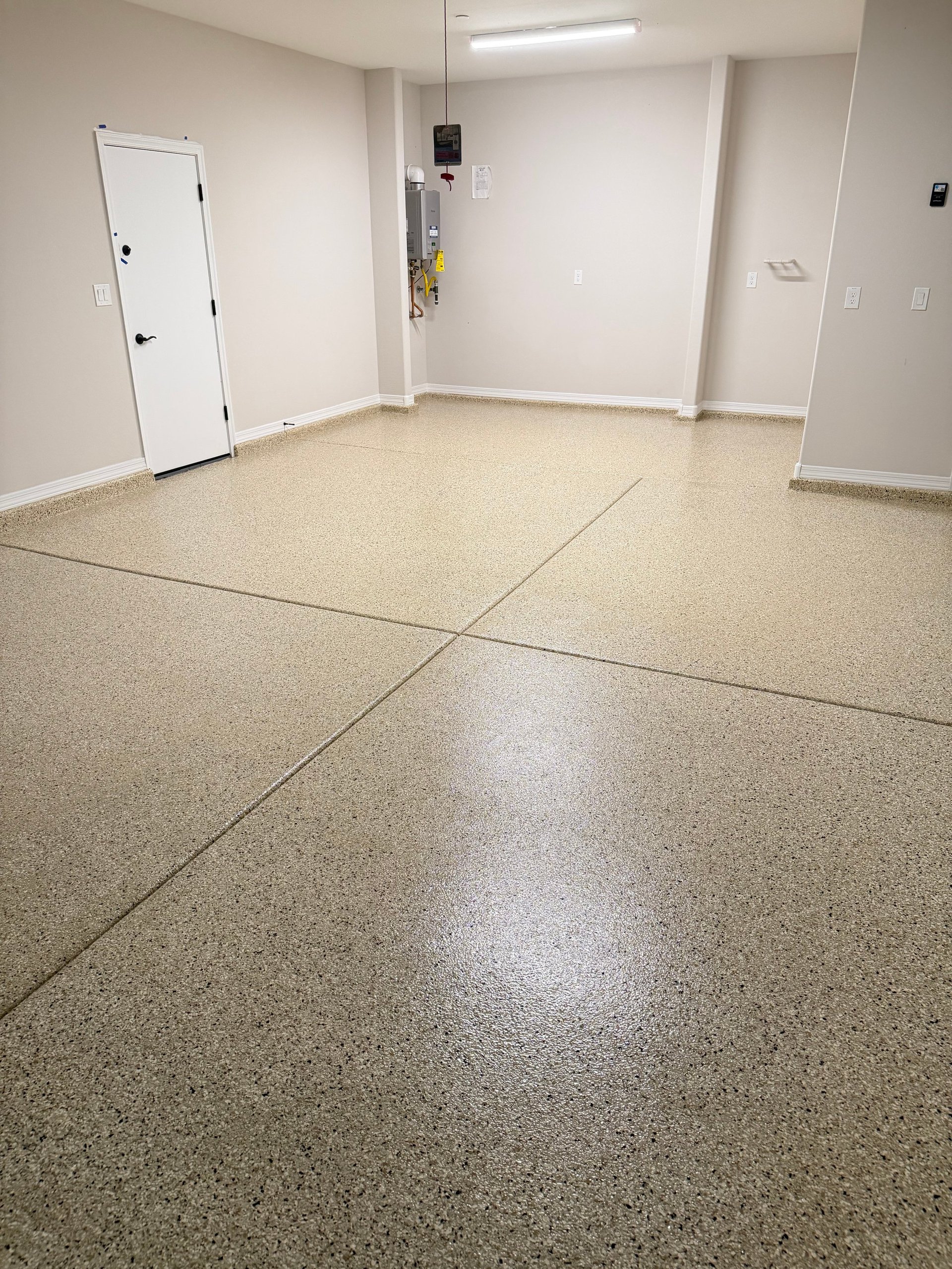 Empty commercial basement room with speckled epoxy flooring, white walls, a white door on the left, and fluorescent lighting overhead