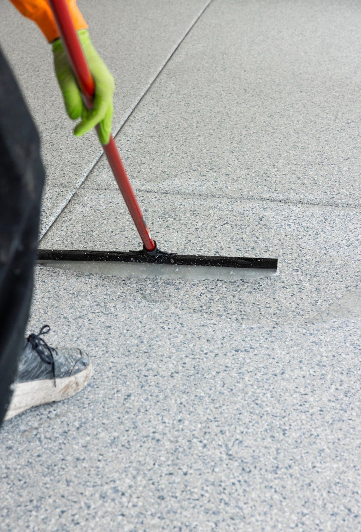 Person wearing green gloves using a squeegee to apply dark sealant or grout between concrete pavement tiles