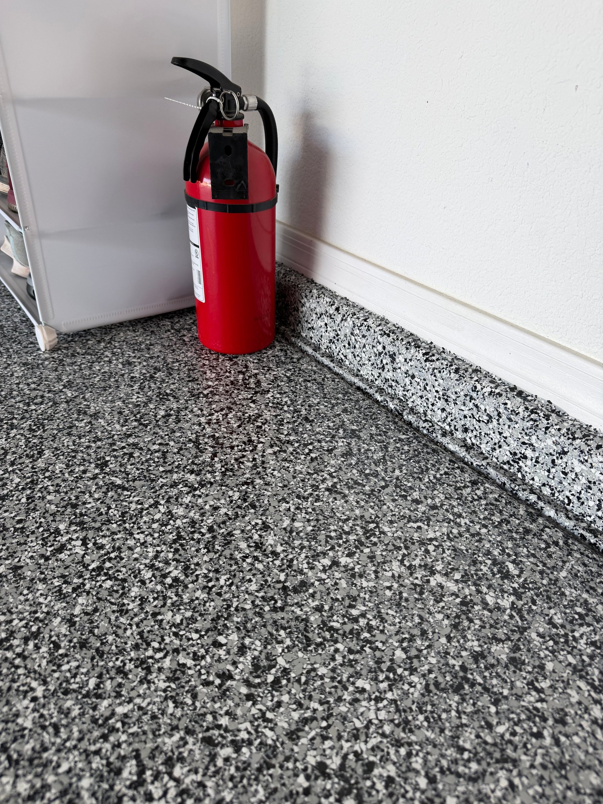 Red fire extinguisher positioned on gray speckled floor against white wall in hallway